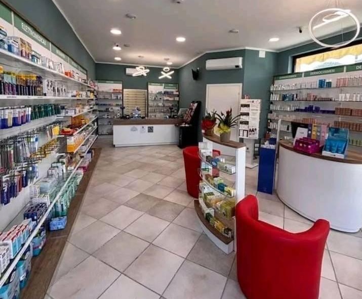 Interno di farmacia con scaffali bianchi e pareti verdi, scritte Elettromedicali e Dermocosmesi, poltroncine rosse e banconi