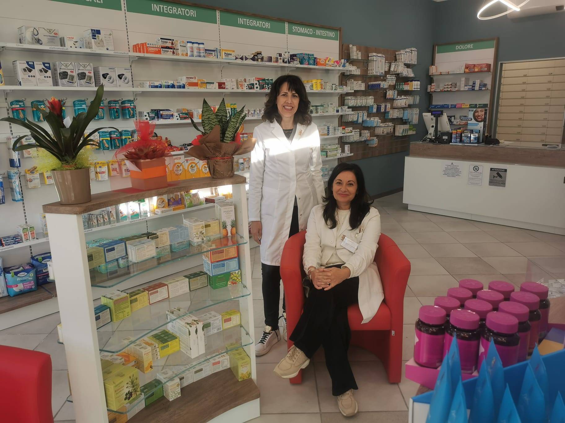 Due farmaciste in camice bianco, una seduta su poltrona rossa, tra scaffali di integratori e piante nella Farmacia Centenario